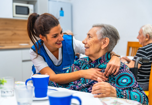 caregiver interacting with elderly woman smiling in a communal dining area senior care support